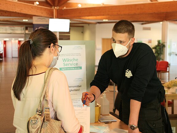 Eine Person im Gespräch mit einem Mitarbeiter am Empfang. Der Mitarbeiter trägt eine Maske und hält einen Stift in der Hand, während er auf ein Formular zeigt. Im Hintergrund steht ein Schild mit Informationen zum Wäscheservice, der täglich angeboten wird.
