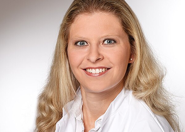 Porträtfoto: lächelnde erwachsene Frau mit langen, welligen, blonden Haaren. Sie blickt direkt in die Kamera. Neutraler, hellgrauer Studiohintergrund. Sie trägt weiße Oberbekleidung und kleine Ohrstecker.