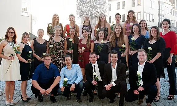 Gruppenfoto: Eine große Gruppe junger Erwachsener in festlicher Kleidung posiert draußen. Viele halten weiße Rosen. Sie lächeln in die Kamera, stehen und knien in Reihen – wirkt wie Abschluss- oder Ehrungsfeier.