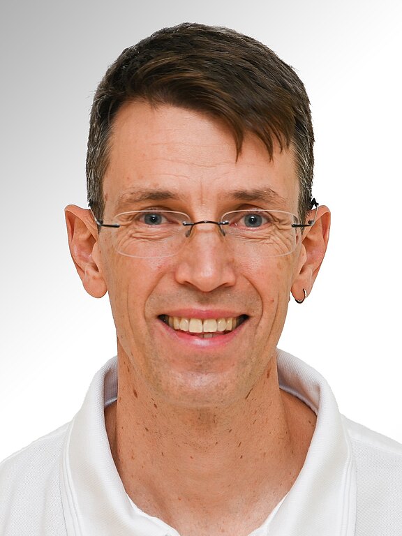 Porträt einer lächelnden Person mit kurzbraunem Haar und schmaler Brille. Sie trägt ein weißes Oberteil und einen kleinen Ohrring am linken Ohr. Neutraler, heller Hintergrund; Fokus auf dem freundlichen Gesichtsausdruck.