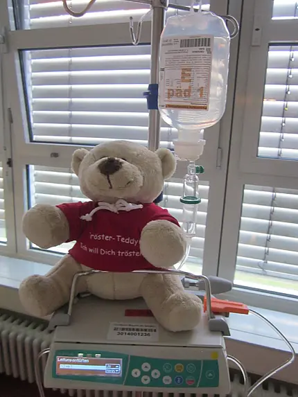 Ein Plüschbär mit rotem T‑Shirt „Tröster‑Teddy“ sitzt auf einer Infusionspumpe. Über ihm hängt ein Beutel mit Infusionslösung am Ständer. Krankenhausumgebung; das Kuscheltier soll Patientinnen und Patienten Trost spenden.