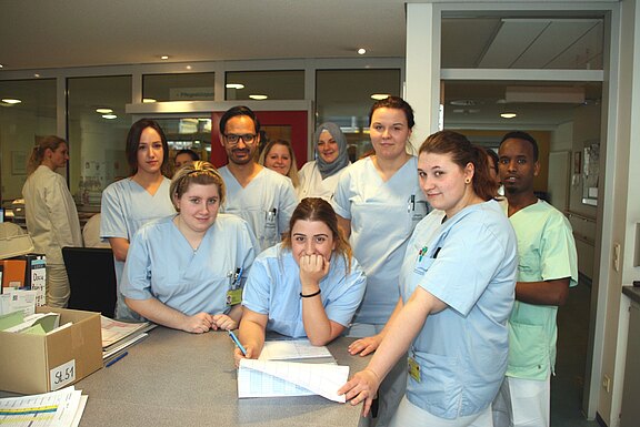 Gruppe von Krankenhausmitarbeitenden in hellen Kitteln steht an einer Stations-Theke. Einige lächeln in die Kamera, eine Person stützt sich über Akten mit Stift. Eindruck: Team von Pflegekräften bei Übergabe oder kurzer Besprechung.