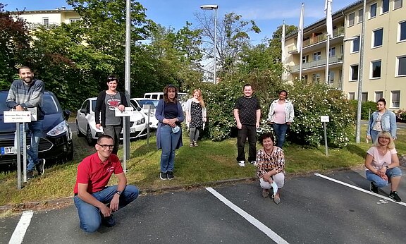 Eine gemischte Gruppe von zehn Erwachsenen posiert im Freien auf einem Parkplatz vor Büschen und einem Gebäude. Einige knien vorn, andere stehen mit Abstand. Lockeres, freundliches Teamfoto; einzelne halten Schutzmasken in der Hand.