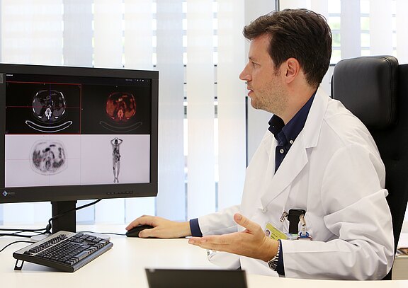 Ein Arzt in einem weißen Kittel sitzt an einem Schreibtisch und betrachtet mehrere medizinische Bilder auf einem Bildschirm. Er gestikuliert während eines Gesprächs, möglicherweise über diagnostische Ergebnisse. Der Raum hat Fenster mit vertikalen Jalousien, die das Licht filtern.