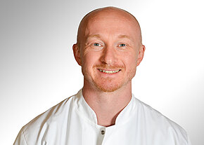 Dr. med. Fabian Hüttl Porträt eines lächelnden, glatzköpfigen Mannes mit kurzem Bart in weißem medizinischem Kittel vor hellem, neutralem Hintergrund; direkter Blick in die Kamera.