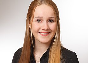 Kristin Beier Porträtfoto: junge Frau mit langen, glatten, hellblonden Haaren, lächelt freundlich. Sie trägt eine schwarze Bluse und eine zarte Halskette vor neutralem, hellem Hintergrund.