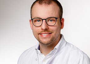 Porträt eines lächelnden Mannes mit Brille, kurzem Haar und leichtem Bart, der einen weißen Kittel trägt. Neutraler Hintergrund.