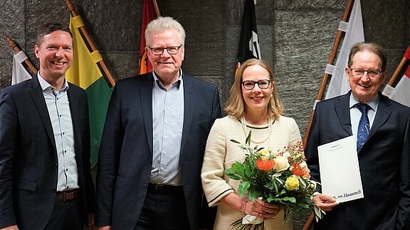 Eine Gruppe von vier Personen steht lächelnd zusammen. Eine Frau hält einen Blumenstrauß in der Hand, während einer der Männer ein Dokument präsentiert. Im Hintergrund sind mehrere Fahnen sichtbar. Die Atmosphäre wirkt feierlich und freundlich.