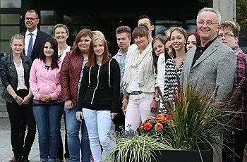Eine Gruppe von Jugendlichen steht in einem Außenbereich, umgeben von Pflanzen. Einige von ihnen lächeln, während mehrere Erwachsene, darunter ein Mann in einem Anzug, ebenfalls dabei sind. Die Szenerie vermittelt eine freundliche und einladende Atmosphäre.