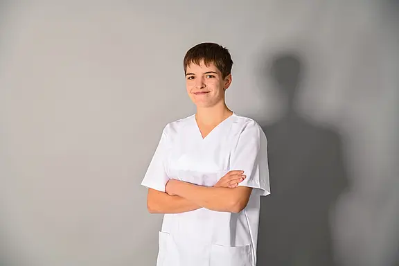 Junge erwachsene Person in weißer Pflegeuniform steht mit verschränkten Armen und lächelt selbstbewusst vor neutralem grauem Hintergrund.