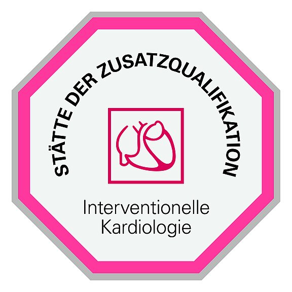 Das Bild zeigt ein oktagonales Schild mit dem Text „Stätte der Zusatzqualifikation“ und „Interventionelle Kardiologie“. In der Mitte befindet sich eine rote Grafik eines Herzens.