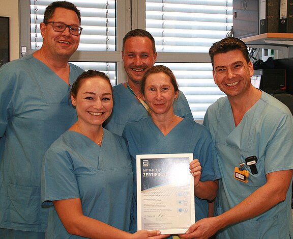 Fünf medizinische Fachkräfte in blauer OP-Kleidung lächeln. Zwei vorn halten ein Zertifikat in die Kamera, das eine Auszeichnung oder erfolgreiche Teilnahme zeigt. Stimmung: stolz und positiv.