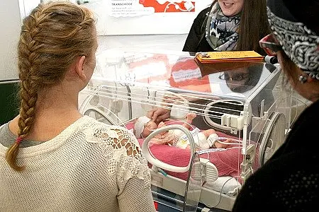 Eine Person mit geflochtenem Haar blickt auf einen Inkubator, in dem ein Neugeborenes liegt. Eine andere Person steht daneben und zeigt auf das Baby. Es herrscht eine freundliche Atmosphäre, während die beiden Personen sich über das Baby austauschen.