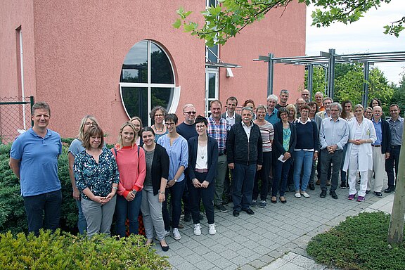 Etwa 30 erwachsene Personen stehen draußen vor einem rosafarbenen Gebäude und posieren für ein Gruppenfoto. Sie lächeln in die Kamera; gemischte Freizeit- und Arbeitskleidung, eine Frau im weißen Arztkittel.