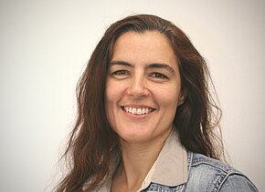 Porträt einer lächelnden Frau mit langen dunkelbraunen Haaren, nach links blickend. Sie trägt eine offene Jeansjacke über einem hellen Hemd. Neutraler, heller Hintergrund. Freundlicher, natürlicher Ausdruck.