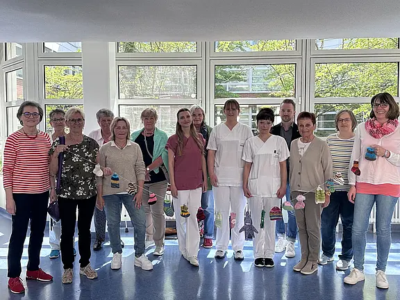 Eine Gruppe von zwölf Personen steht in einem gut beleuchteten Raum mit großen Fenstern. Die Mehrheit trägt lässige Kleidung, einige in Weiß. Sie halten kleine handgefertigte Objekte in den Händen und stehen vor einem Mobile, das aus ähnlichen Objekten besteht.