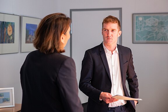 Zwei Personen in Geschäftskleidung führen ein Gespräch in einem Büro. Der Mann hält eine Mappe oder Dokumente in der Hand und schaut die andere Person an. Die Szene wirkt wie ein formelles Meeting oder ein Bewerbungsgespräch.