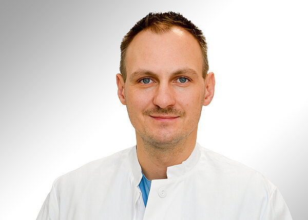 Porträt einer Person in weißem medizinischen Kittel vor hellem, neutralem Hintergrund; Kopf und Oberkörper frontal, freundlicher Gesichtsausdruck.