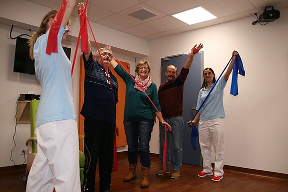 Fünf Erwachsene stehen in einem Therapieraum und machen Gymnastik mit roten und blauen Widerstandsbändern. Zwei Personen in Pflegekleidung führen die Übung an den Seiten an; drei Teilnehmende in Alltagskleidung heben die Arme und spannen die Bänder. Gruppen-Rehabilitation bzw. Bewegungstraining.