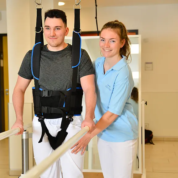 Mann trainiert zwischen Parallelstangen das Gehen, gesichert in einem hängenden Sicherheitsgeschirr. Eine Therapeutin steht daneben und unterstützt seine Schritte. Szene in einer Therapiepraxis.