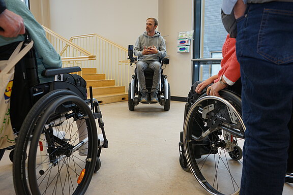In einem hellen Raum spricht ein Mann im Elektrorollstuhl zu einer kleinen Gruppe von Menschen im Rollstuhl. Sie stehen im Halbkreis und hören zu. Im Hintergrund ist eine Treppe sichtbar, Thema scheint Barrierefreiheit oder Erfahrungsaustausch.