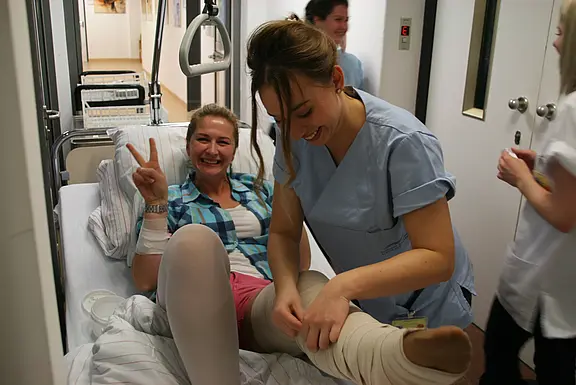 Patientin liegt lächelnd im Krankenhausbett und zeigt das Victory-Zeichen, während eine Pflegekraft ihr am Bein einen Verband anlegt. Die Szene vermittelt medizinische Versorgung und eine positive, unterstützende Stimmung.
