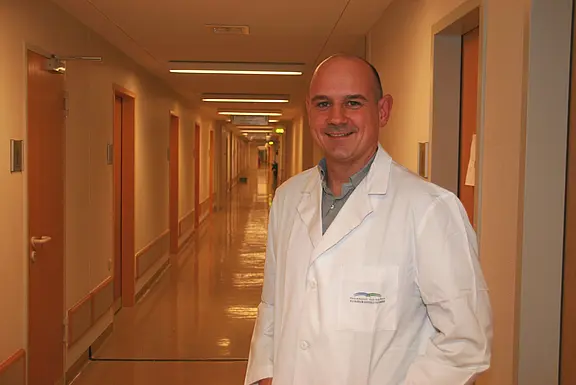 Ein lächelnder Arzt im weißen Kittel steht in einem hellen Krankenhausflur und blickt in die Kamera; das Bild vermittelt eine professionelle, freundliche medizinische Umgebung.