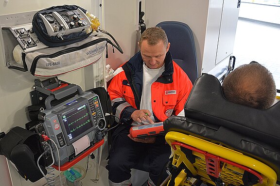 Sanitäter in einem Rettungswagen betreut einen Patienten auf der Trage. Ein Monitor zeigt EKG‑Kurven und Vitalwerte, daneben medizinische Ausrüstung. Der Sanitäter dokumentiert oder bedient ein Gerät. Szene vermittelt Notfallversorgung im Fahrzeug.