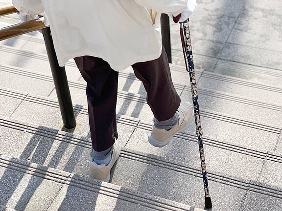 Eine Person mit einer weißen Jacke und dunklen Hosen geht eine Treppe hinunter, während sie sich mit einem blumigen Gehstock abstützt. Ihre Schuhe sind hell und sie hat graue Socken an. Die Stufen sind mit Rillen versehen.