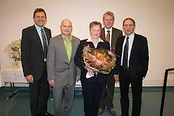 Fünf Personen stehen zusammen, während eine Frau einen großen Blumenstrauß in den Händen hält. Die Männer sind formell gekleidet und lächeln, während sie der Frau gratulieren. Der Hintergrund ist hell und neutral.