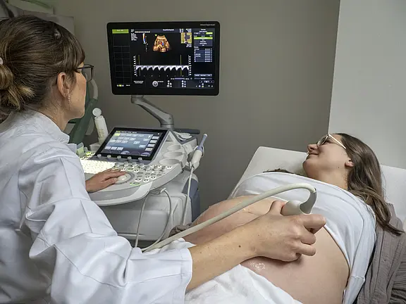 Eine schwangere Frau liegt auf einer Liege und lächelt, während eine Ärztin ein Ultraschallgerät bedient. Der Fokus liegt auf dem Bauch der Frau, wobei ein Ultraschallbild auf dem Bildschirm zu sehen ist. Die Atmosphäre ist beruhigend und informativ.