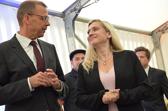 Zwei Menschen in Anzug und Blazer führen ein Gespräch in einem hellen Raum; die Frau lächelt den Mann an. Im Hintergrund stehen zwei weitere Personen.