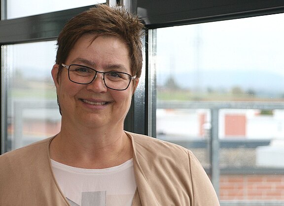 Porträtfoto einer lächelnden erwachsenen Person mit kurzer brauner Frisur und Brille in heller Kleidung. Sie steht in einem modernen Innenraum vor großen Fenstern; der Hintergrund ist unscharf. Der Fokus liegt auf dem freundlichen Gesichtsausdruck.