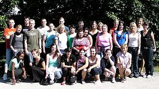 Eine Gruppe von etwa 30 Personen steht in einem Außenbereich, teilweise sitzend und teilweise stehend, vor einer grünen Hintergrundlandschaft. Die Personen sind unterschiedlich gekleidet und scheinen lächelnd in die Kamera zu blicken.