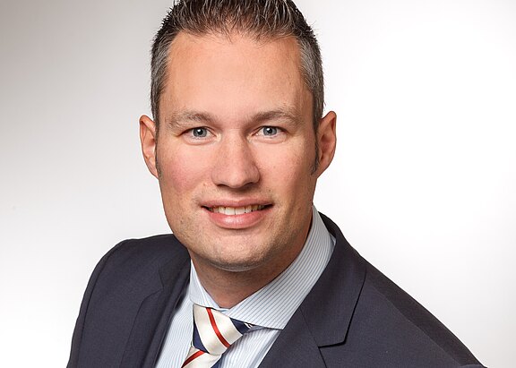 Porträt eines Mannes mit kurzen, grauen Haaren und einem freundlichen Lächeln. Er trägt einen dunklen Anzug und ein gestreiftes Hemd mit Krawatte. Der Hintergrund ist hell und einfarbig, um die Person hervorzuheben.