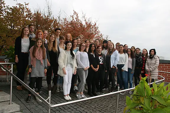 Große Gruppe junger Menschen steht dicht beisammen und lächelt in die Kamera, als Gruppenfoto im Freien. Sie posieren auf einem gepflasterten Weg mit Geländer; im Hintergrund herbstlich gefärbte Bäume. Fokus auf die gesamte Gruppe.