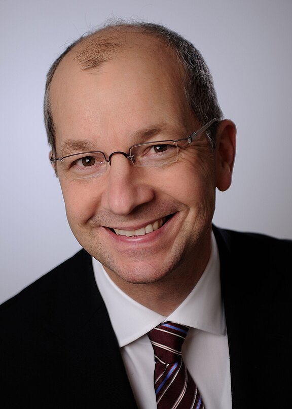 Porträtfoto eines lächelnden, mittelalten Mannes mit Brille. Er trägt dunklen Anzug, weißes Hemd und gestreifte Krawatte. Neutraler heller Hintergrund, Kopf- und Schulteraufnahme, freundlicher Blick direkt in die Kamera.