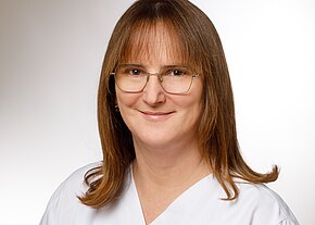 Station 33A Porträt einer lächelnden erwachsenen Person mit schulterlangem braunem Haar und Brille, frontal vor neutralem Hintergrund. Sie trägt weiße Arbeitskleidung.