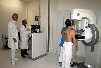 Ein Kind steht vor einem medizinischen Gerät zur Untersuchung, während ein Arzt daneben am Computer arbeitet. Eine Person in blauer Kleidung assistiert dem Kind, das sich für die Untersuchung vorbereitet. Im Hintergrund sind weitere Fachleute zu sehen.