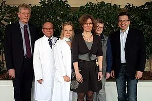 Sechs Personen stehen zusammen vor einer grünen Pflanze. Drei Personen tragen formelle Kleidung, während eine Person einen weißen Laborkittel trägt. Die anderen beiden Personen sind in Alltagskleidung. Die Gruppe scheint sich in einem freundlichen und professionellen Umfeld zu befinden.