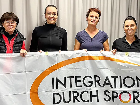 Fünf Frauen halten gemeinsam ein großes Banner mit der Aufschrift „Integration durch Sport“. Sie stehen nebeneinander und lächeln; die Botschaft betont Zusammenhalt und Teilhabe durch Sport.