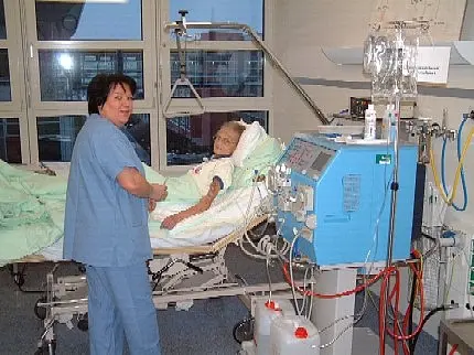 Patientin bei der Hämodialysebehandlung Pflegekraft betreut eine liegende Person im Krankenhausbett. Neben dem Bett steht eine große medizinische Maschine mit Schläuchen und Monitor; die Person ist daran angeschlossen.