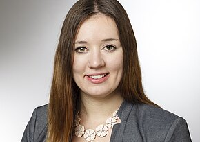 Sandra Bergmann Portrait einer lächelnden Frau mit langen, braunen Haaren und einem grauen Blazer. Sie trägt eine auffällige Halskette mit blütenförmigen Anhängern. Der Hintergrund ist hell und einfarbig, was den Fokus auf ihr Gesicht lenkt.