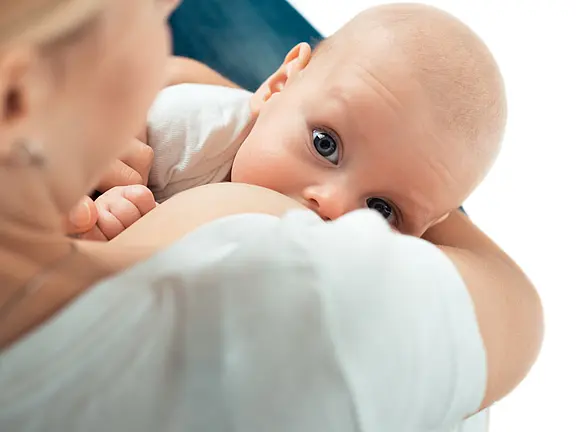 Nahaufnahme: Ein Säugling trinkt an der Brust seiner Mutter und blickt nach oben. Der Kopf liegt im Arm, ruhige Szene, Fokus auf Stillen und Bindung.