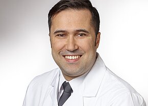 MD Fadi Alhalaby Ein lächelnder männlicher Arzt trägt einen weißen Kittel und eine Krawatte. Der Hintergrund ist hell und einfarbig. Das Bild vermittelt Freundlichkeit und Professionalität.