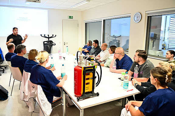 Ein Vortrag findet in einem Schulungsraum statt. Ein Referent steht vorne und erklärt Inhalte an einem Bildschirm. Eine Gruppe von Fachleuten in blauen Uniformen sitzt an einem Tisch und hört aufmerksam zu. Verschiedene Trinkflaschen stehen auf dem Tisch.