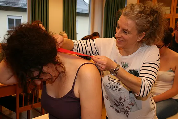Eine lächelnde Frau bringt einer sitzenden Frau einen roten Kinesiologie‑Tape‑Streifen am Nacken und Schulterbereich an, wie in einem Workshop oder Kurs. Fokus auf die praktische Taping‑Anwendung zur Entlastung des Nackens.