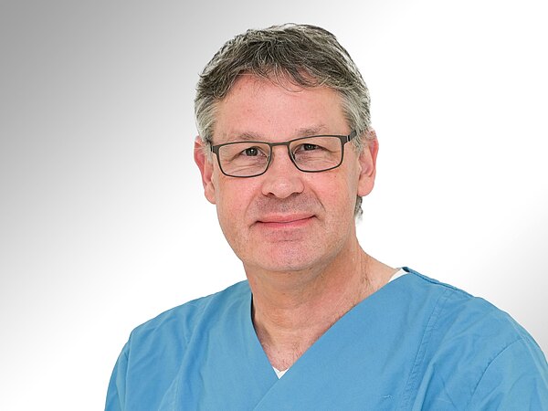 Ein Mann mit grauen Haaren und Brille trägt einen blauen OP-Overall. Er lächelt freundlich in die Kamera. Der Hintergrund ist hell und neutral, was den Fokus auf seine Person legt.