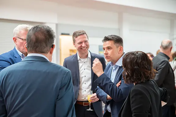 Mehrere Erwachsene in Businesskleidung unterhalten sich lebhaft bei einem Empfang. Ein Mann gestikuliert mit einem Getränk in der Hand, andere hören lächelnd zu. Atmosphäre: berufliches Netzwerkgespräch.