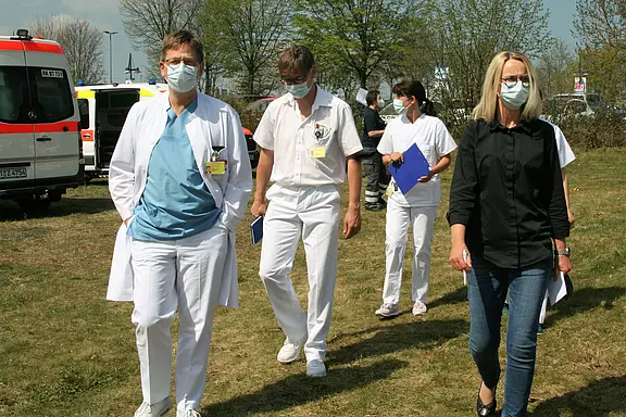 Fünf Personen, darunter medizinisches Personal in weißen Kitteln und einer in einem schwarzen Hemd, gehen auf einem Grasfeld. Alle tragen Gesichtsmasken. Im Hintergrund sind Rettungsfahrzeuge und Bäume zu sehen.